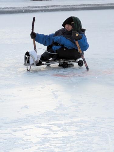 A sled hockey player is demonstrating how to move a hockey sled, wearing a blue coat with hood up to protect from the wind on the outdoor ice rink.