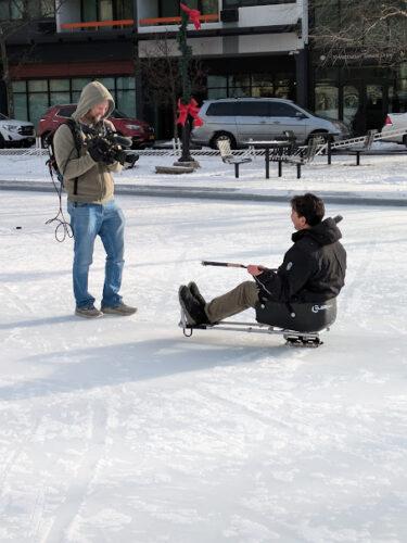 Two people on an outdoor skating rink; one is holding a tv camera trained on the other person who is in a hockey sled.