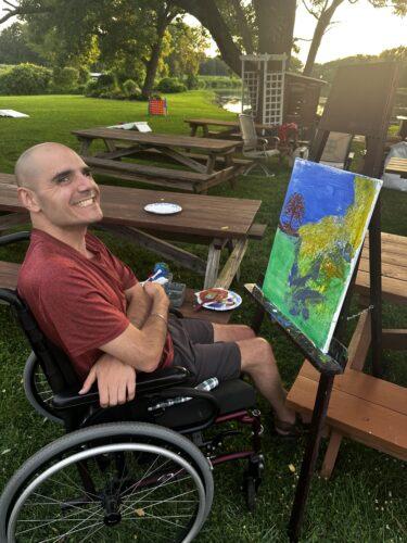 A man with closely shaved hair sits in a manual wheelchair with arms folded across his lap, as he smiles towards the camera; he is seated in front of a canvas with many bright colors on an easel.