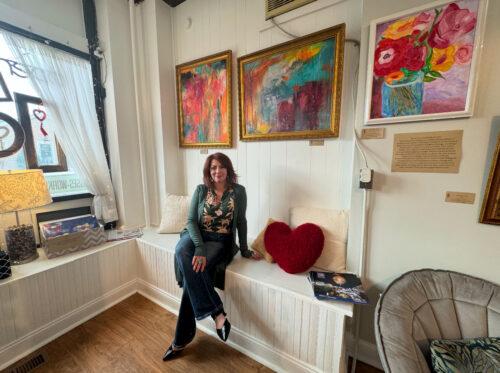 A woman is seated on a wooden built in bench in an art studio, with colorful artwork on the wall behind her.