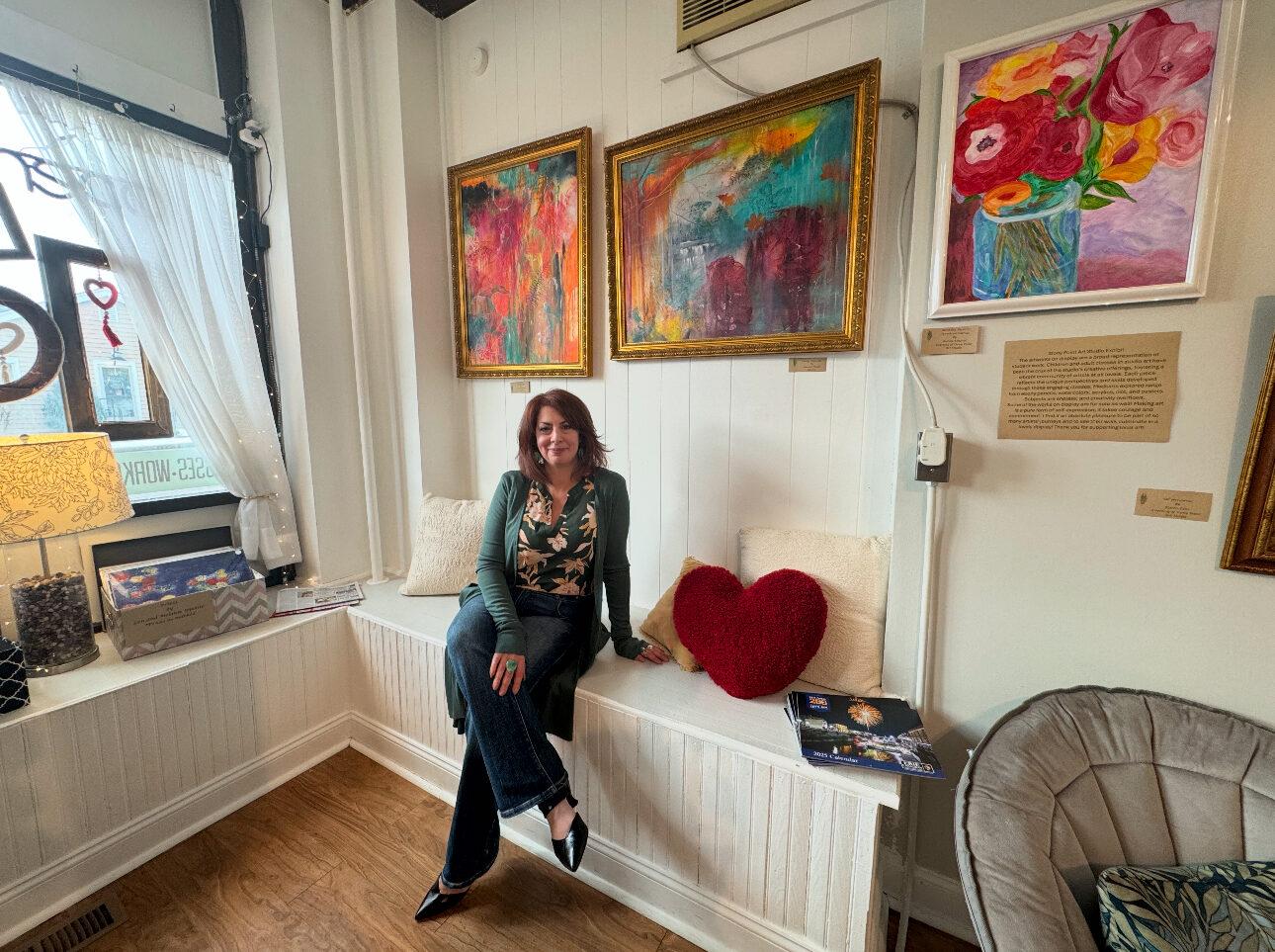 A woman is seated on a wooden built in bench in an art studio, with colorful artwork on the wall behind her.