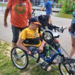 A woman in an orange RAA tshirt stands by a young man who is preparing to ride a handcycle.