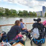 A group of people in manual and power wheelchairs are on the deck of a boat, touring along the Genesee Riverway with trees and a building in the background.