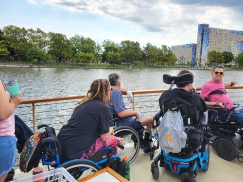 A group of people in manual and power wheelchairs are on the deck of a boat, touring along the Genesee Riverway with trees and a building in the background.