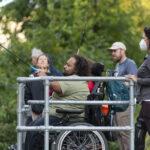 Several people at an accessible fishing rail with green bushes and trees in the background. A man in a manual wheelchair is casting his fishing line in the foreground.