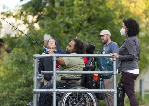 Several people at an accessible fishing rail with green bushes and trees in the background. A man in a manual wheelchair is casting his fishing line in the foreground.