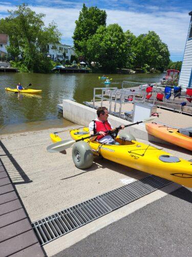 A man sitting in a yellow kayak that is using a kayak chariot device with wide tires to roll up out of the water on a cement rampway.