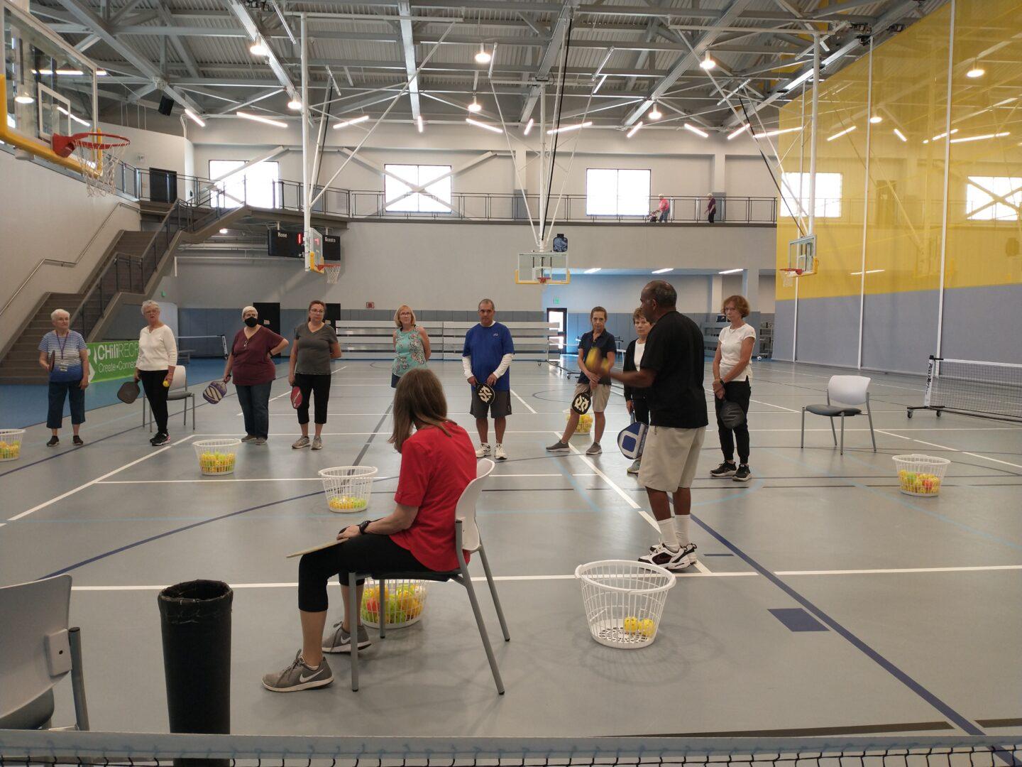 A large gym space with pickleball courts and people standing or sitting near an instructor