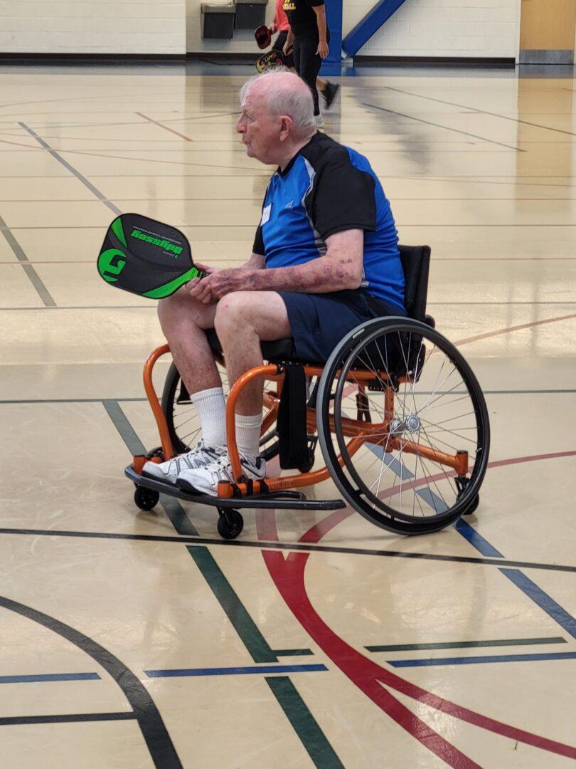 A man in a sports chair on an indoor pickleball court with paddle extended to receive a volley