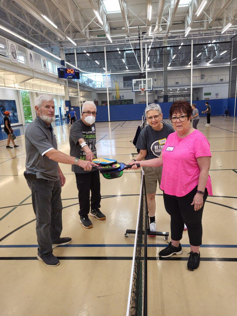 Two people standing on either side of an indoor pickleball net each with their paddles extended to the middle so all paddles are touching
