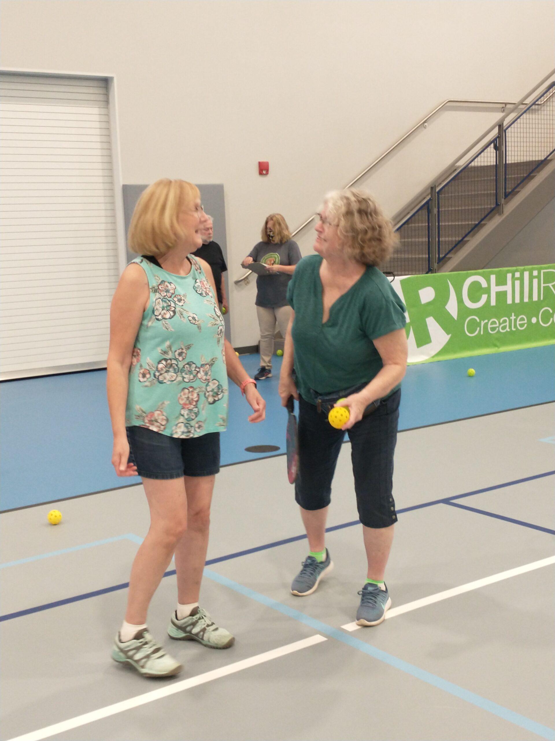 Two women stand on an indoor pickleball court talking, one of them holding a pickleball