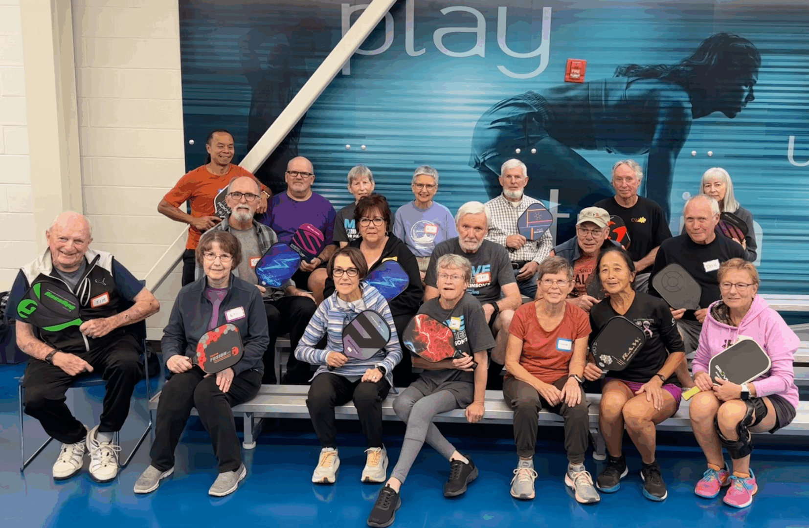 A group of twenty people in three rows of a small bleacher in a gym each holding a pickleball paddle