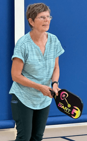 A woman in a light blue shirt stands with her pickleball paddle ready to return a serve