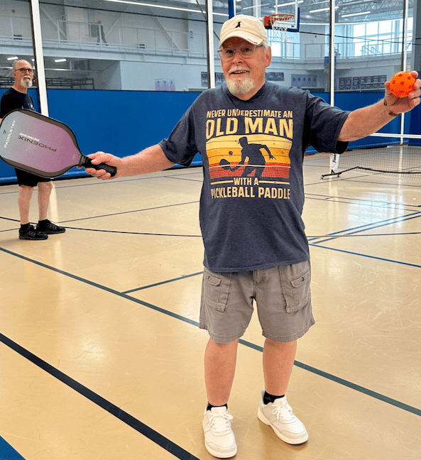 A man with a shirt that reads Never Trust an Old Man with a Pickleball Paddle holds a pickleball paddle and orange ball