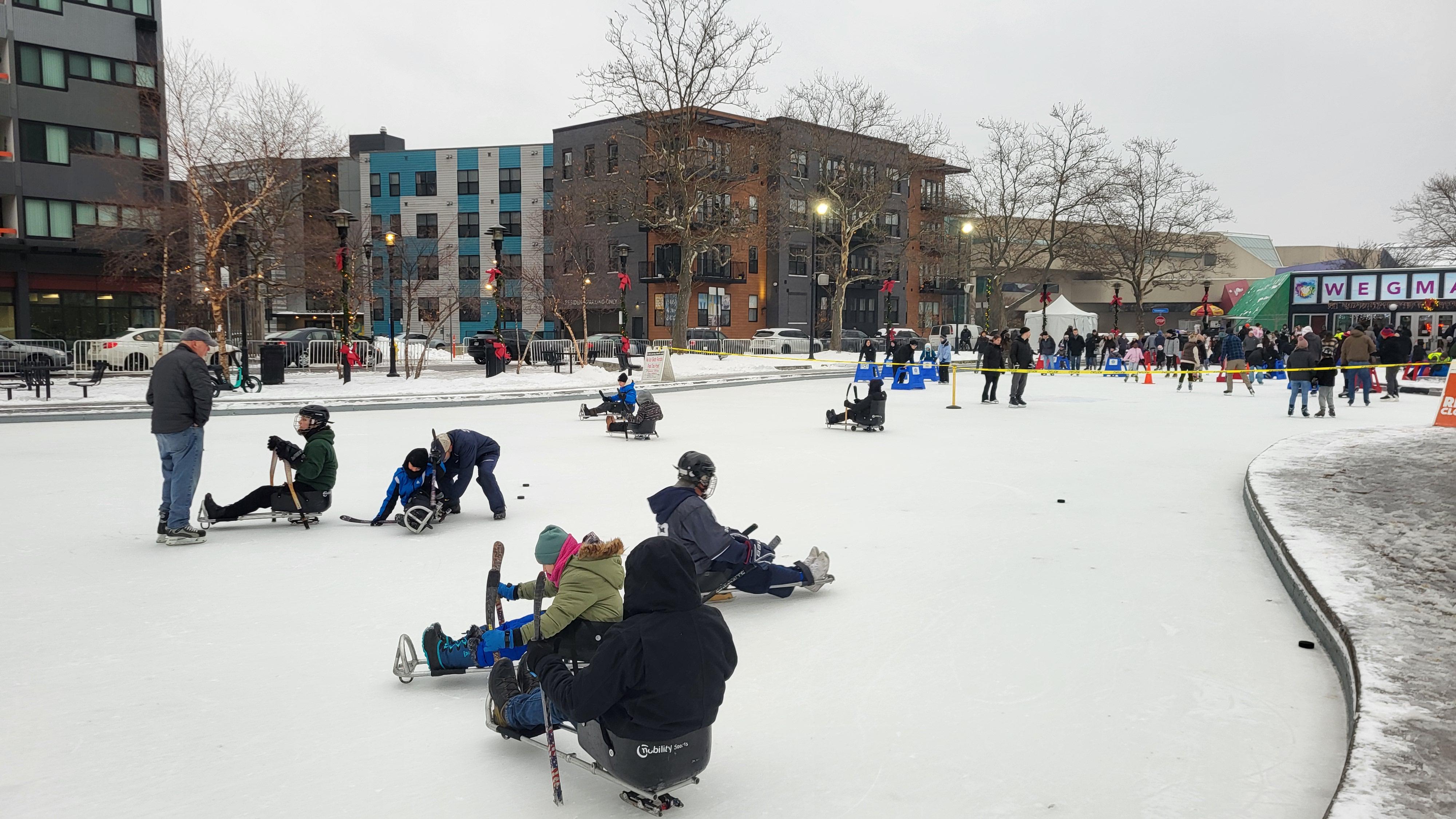 A view across an outdoor ice rink with several people skating using sleds.