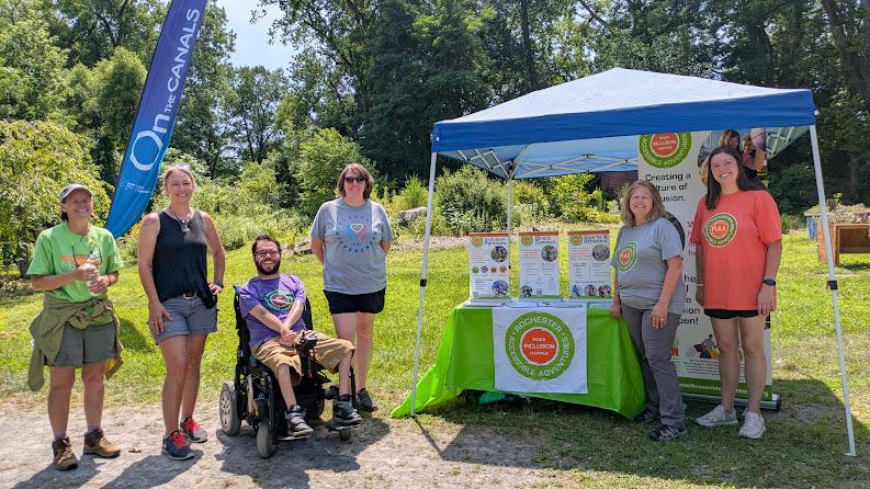 A group of people, one in a power wheelchair and others standing, are outside on a sunny day with grass and trees behind them and an RAA info table and ten