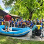 A large group of people, many in wheelchairs or other mobility devices, listen to a rafting instructor prior to going rafting