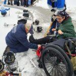 A Sled Hawks player preps her equipment as the coach and volunteer support her shifting from her wheelchair to the hockey sled and then onto the outdoor rink