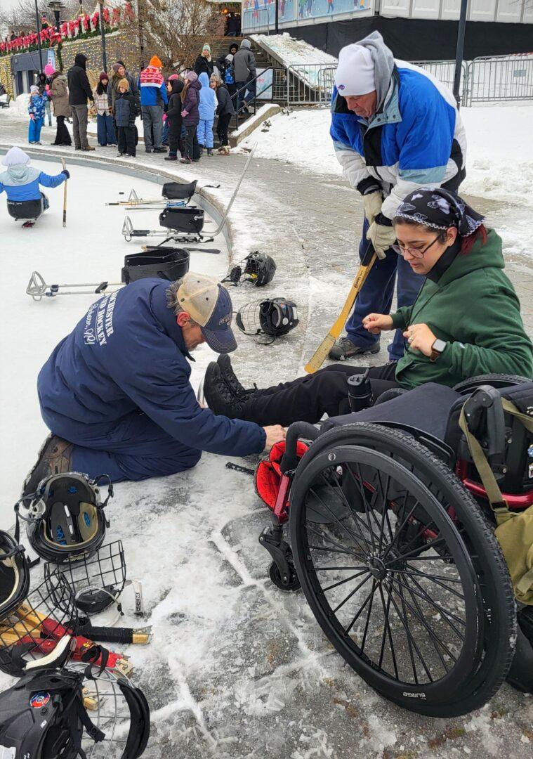A Sled Hawks player preps her equipment as the coach and volunteer support her shifting from her wheelchair to the hockey sled and then onto the outdoor rink