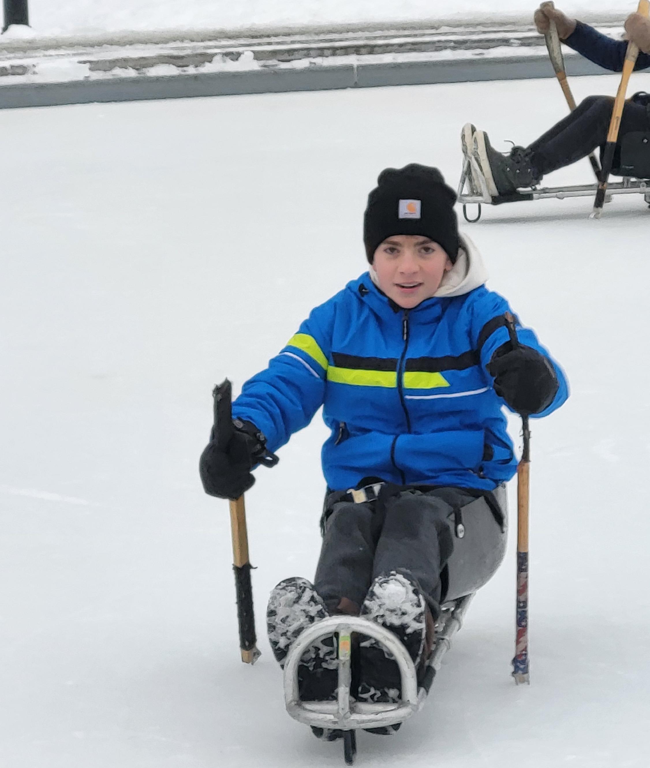 A teenager who is volunteering for RAA tries out sled hockey seated in a sled with sticks on an outdoor ice rink