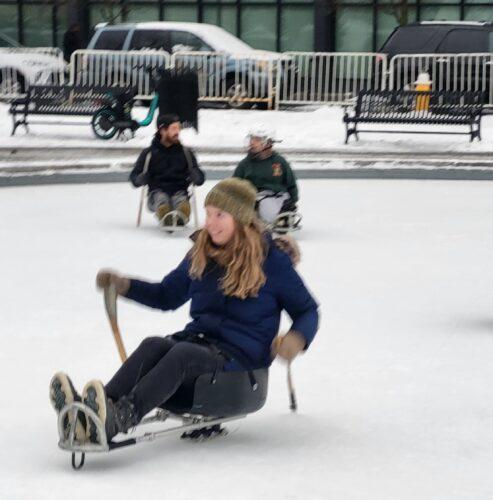 An RAA volunteer tries out sled hockey on an outdoor ice rink and is digging in the sticks to propel her forward