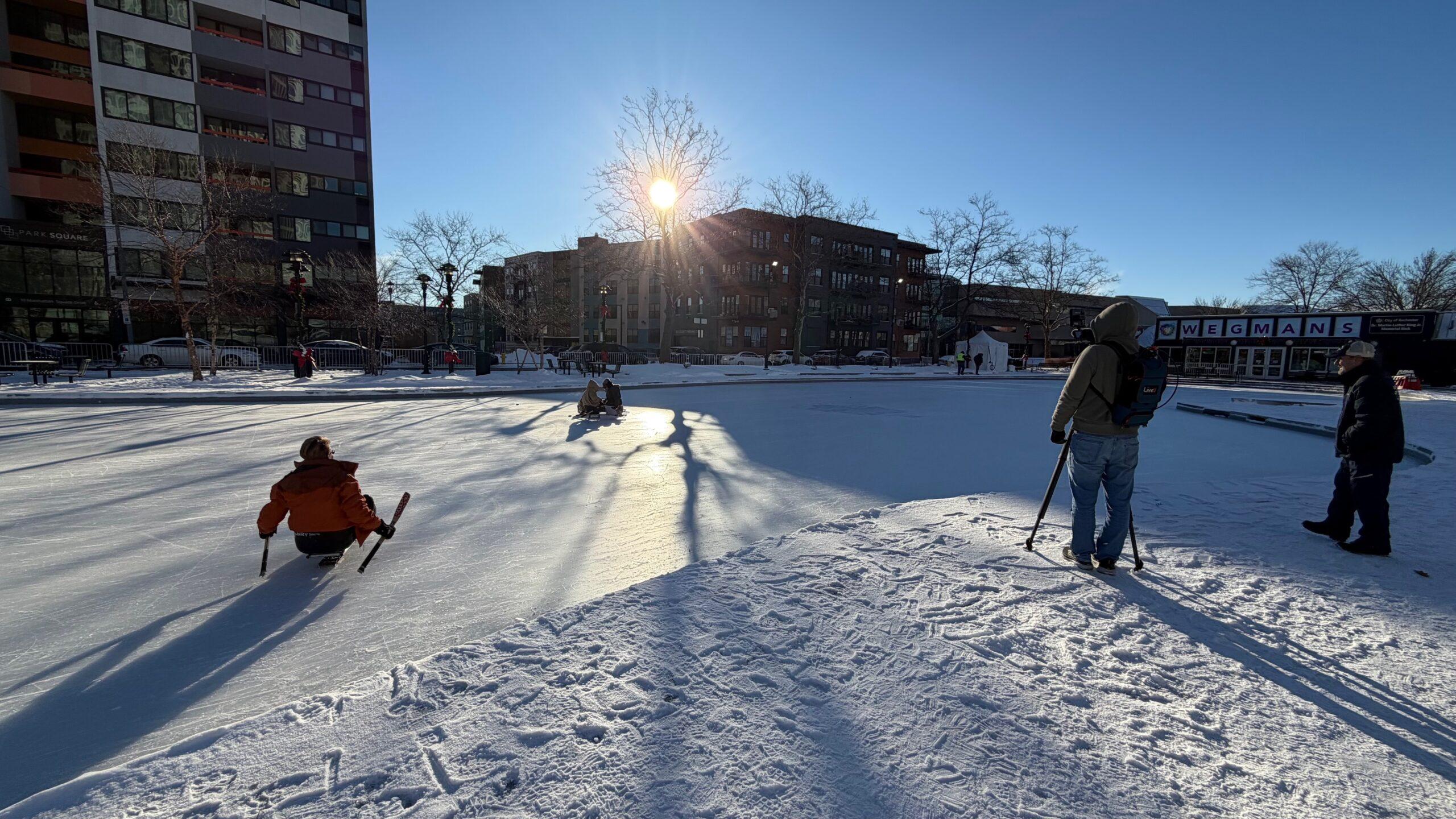 A bright blue sky and city buildings are the background to a dazzling outdoor ice rink where a videographer is filming a reporter trying out sled hockey