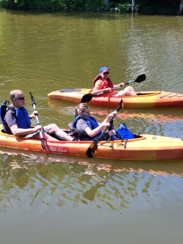 Two family members kayak together in a tandem kayak, the front seat outfitted with adaptive seatback and paddle pedestal. A volunteer paddles nearby.