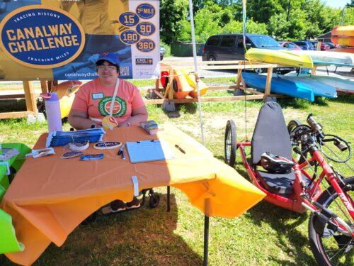 An RAA volunteer is seated behind a table displaying info on the Canalway Challenge at an RAA event; a red handcycle is on the grass beside the table