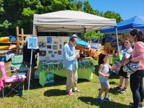 A girl and her mom enjoy conversing with a woman at the GVAS birding tent at an extravaganza with bright sunshine on the grass