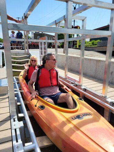 Two people in a orange and yellow tandem kayak on a BoardSafe adaptive kayak launch are prepping to move forward out into the water at the Canal