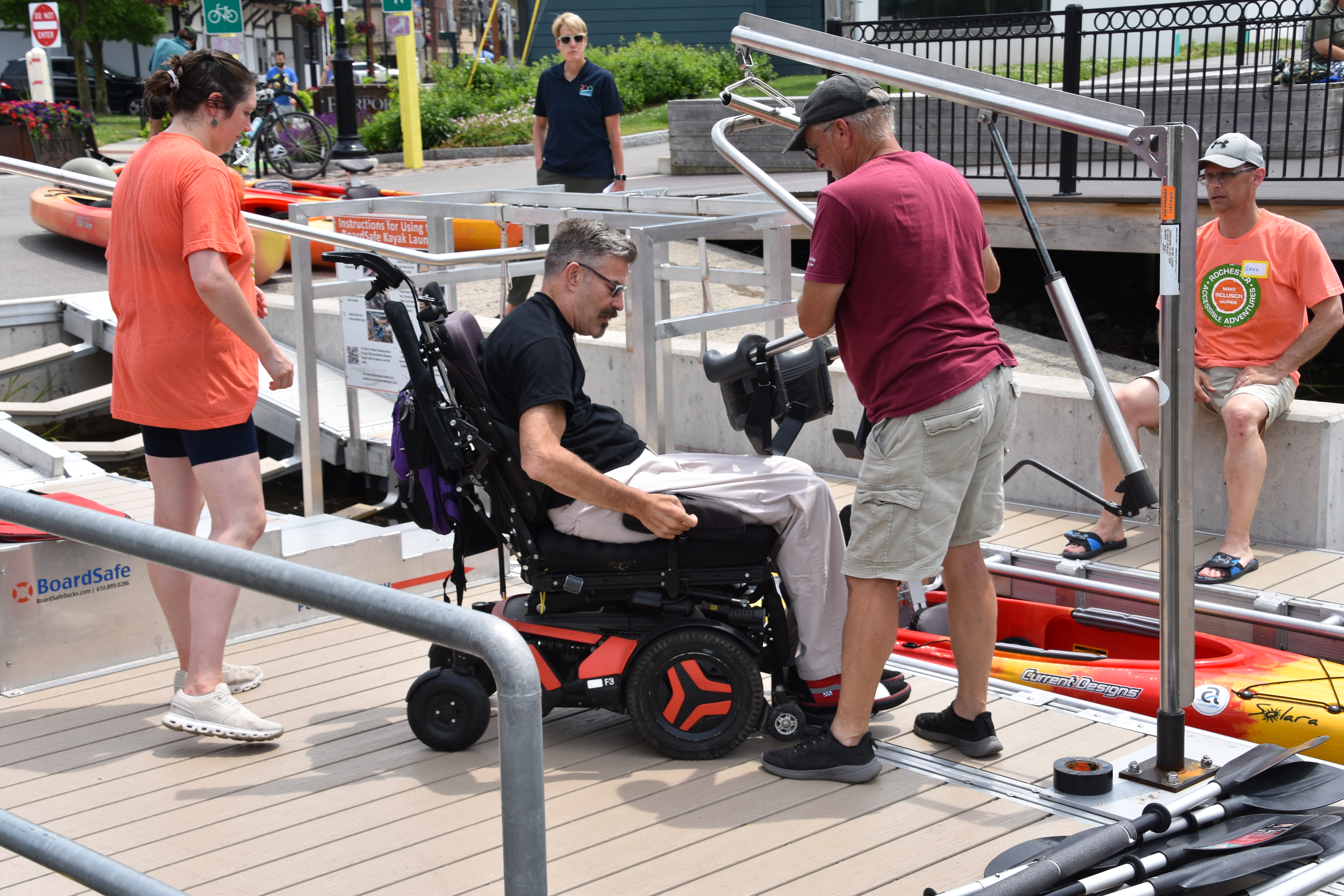 A man who uses a power wheelchair is working with staff to prep for transferring into a kayak that awaits him on the roller-free adaptive launch area