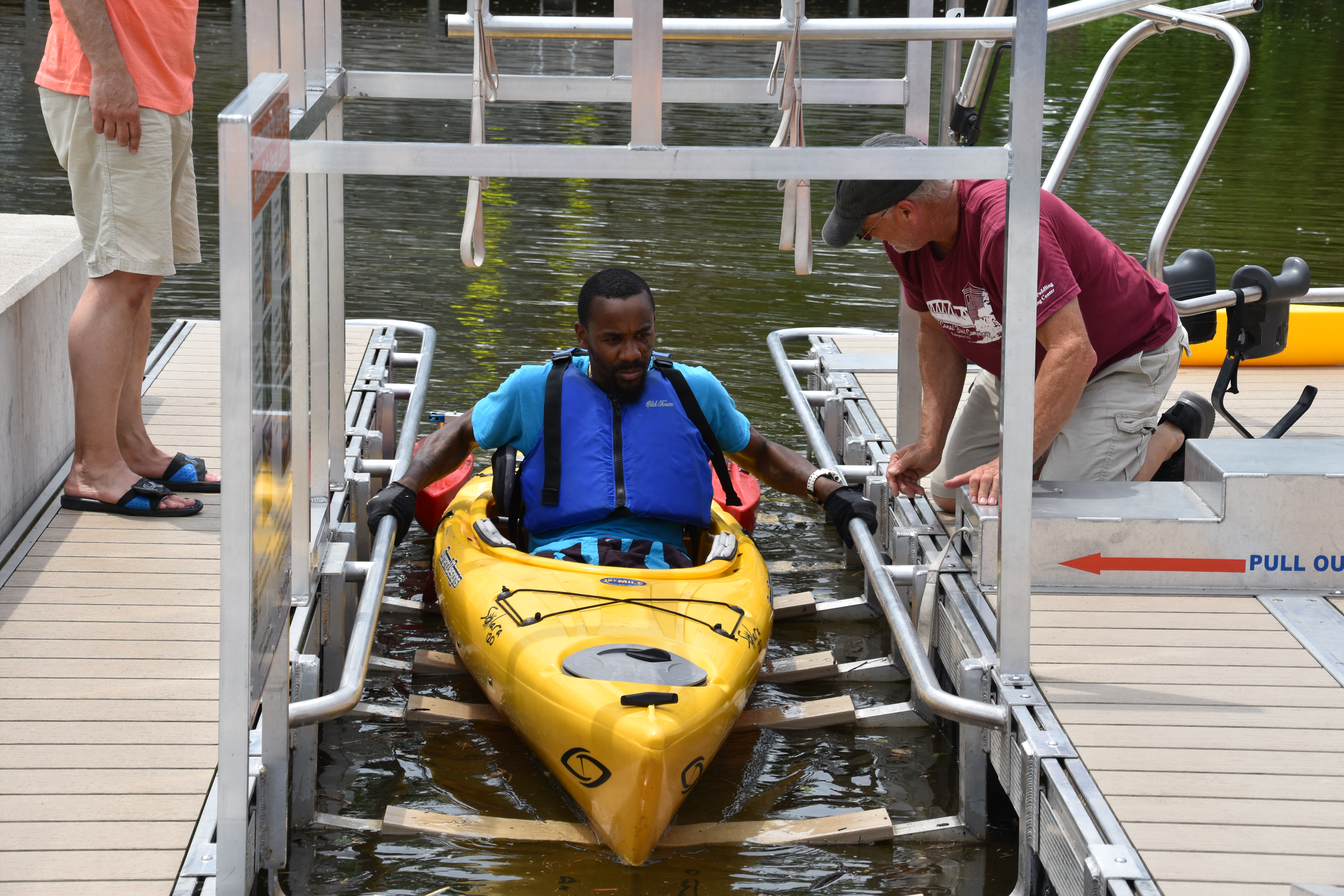 A man in a yellow kayak gets adaptive equipment fitted as he preps to exit the kayak on a BoardSafe adaptive kayak launch that has a system of bars and straps to help exit the boat