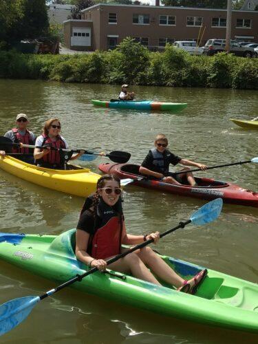 A family kayaks together on the Erie Canal, the parents in a tandem and two siblings each in a single kayak