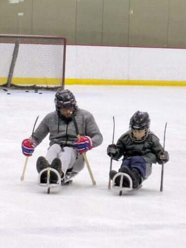 An adult and a child on hockey sleds dig their sticks into the ice to pull forward on an indoor ice rink