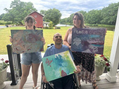 Three participants in an outdoor art class hold up their canvasses which are painted in variations of green and purple pastels. One participant uses a manual wheelchair while two stand on either side