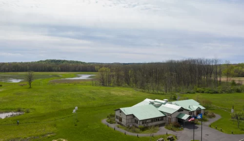 A wide angle view of a nature expanse with a nature center building in the lower right corner and extending out over a green expanse lined by trees