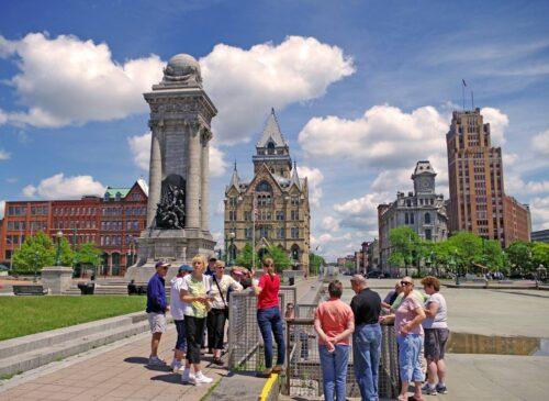 A group of people gather around a speaker during an excursion in Syracuse with tall buildings in the background on a sunny summer day