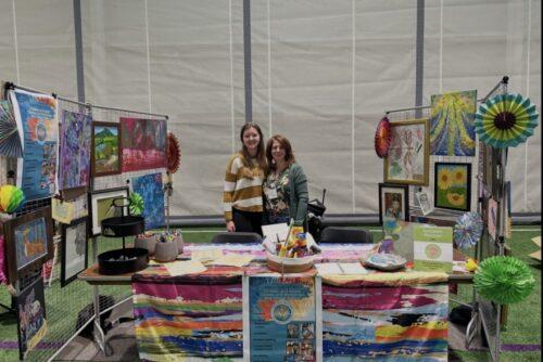 Two women stand behind a table at an indoor festival with a table full of artistic displays and tall stands beside and behind them displaying a variety of art mediums and artwork.