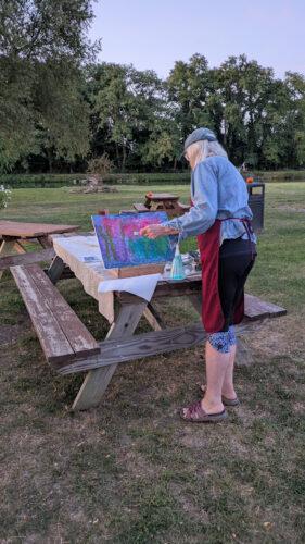 A person stands outdoors by a picnic table painting on a raised canvas