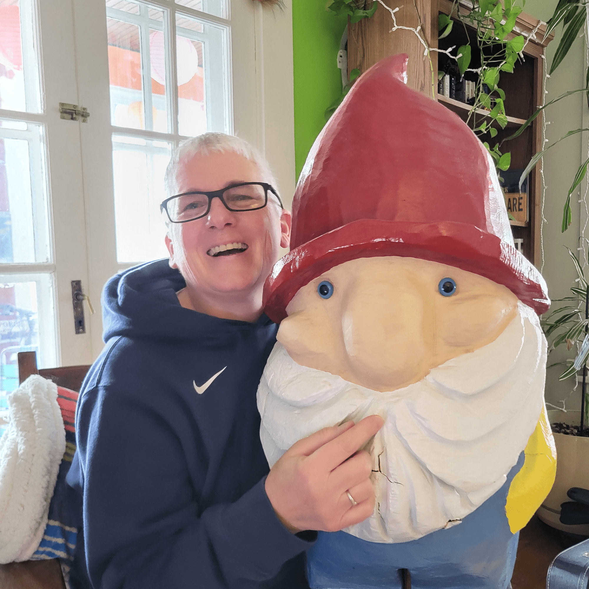 A woman with short white hair and glasses is laughing beside a wooden gnome with red hat and white beard and blue pants
