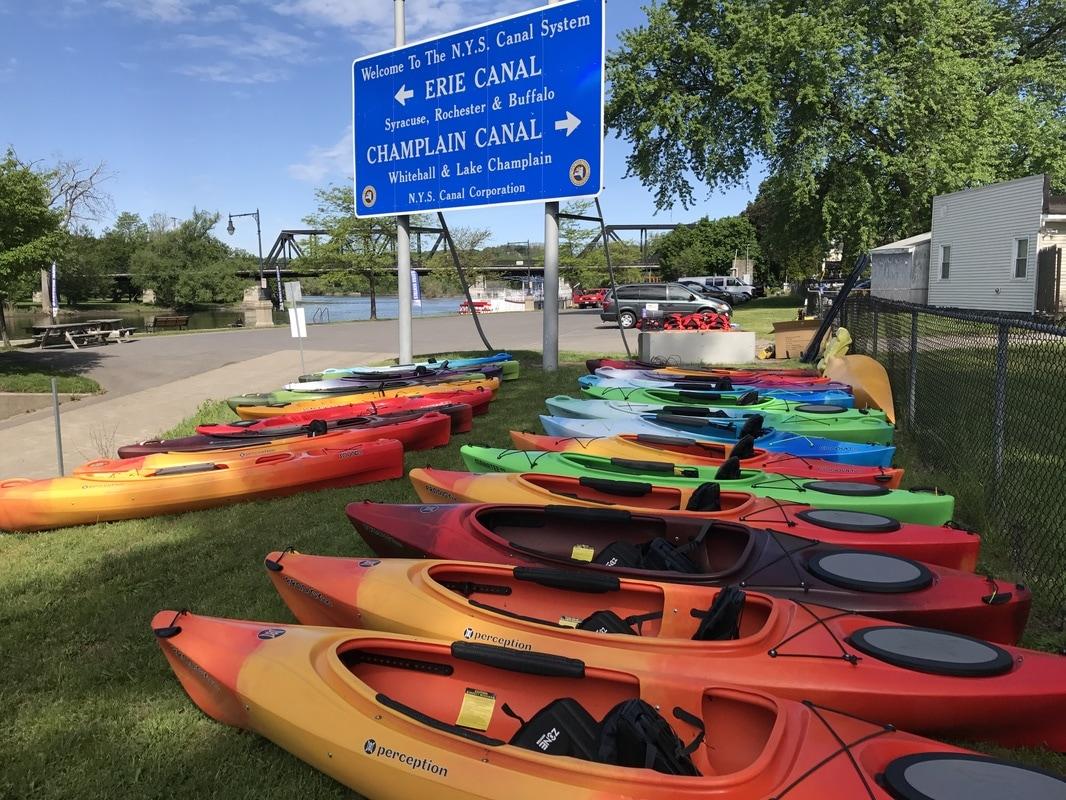 A row of colorful kayaks on a grassy area beside a large blue sign heralding the Erie Canal and Champlain Canal