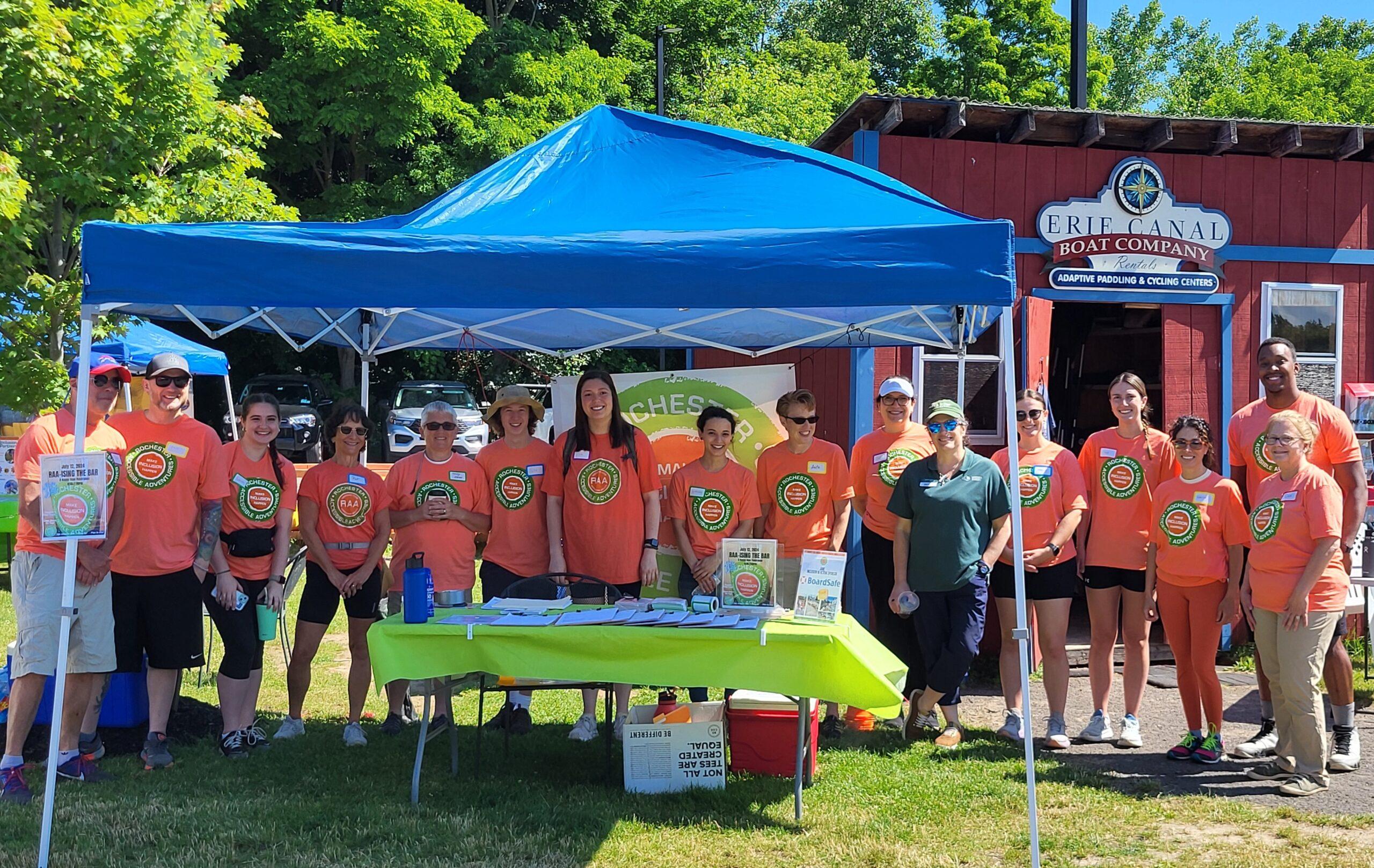 A group of more than 10 people mostly in bright orange RAA shirts stand under and to the side of a blue event tent a Erie Canal Boat Company ready for the ExtRAAvaganza