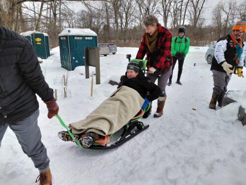 A man in a black coat and winter hat is tucked into an adaptive sled with blankets and one person is pulling the rope on the sled and another is pushing from behind.