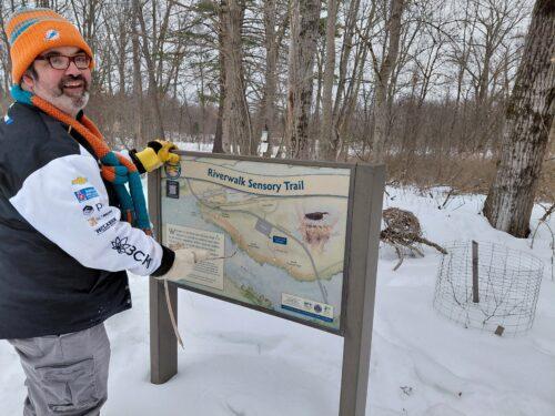 A man in winter coat, hat and gloves stand beside HCP's Riverwalk Sensory Trail sign before heading out on the snow covered trail