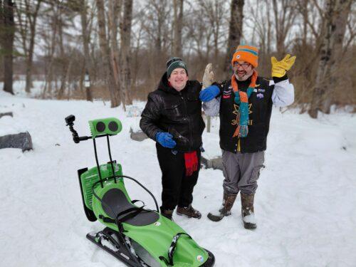 Two men joyfully smiling at a park in the snow with wooded area behind them and a green adaptive sled on the ground beside hem