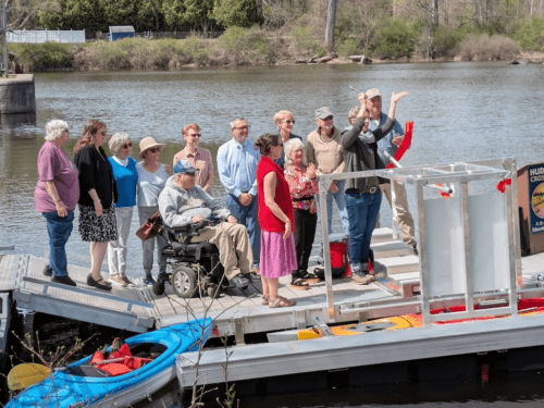 A group of people cheer as a ribbon is cut across a BoardSafe Kayak launch in Hudson Crossing Park.