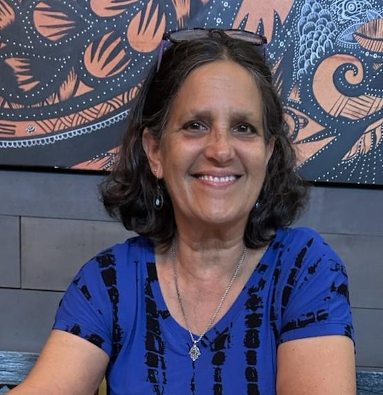 A woman with dark brown shoulder length hair parted in the middle, wearing a blue and black top, a big smile and her glasses perched on top of her head