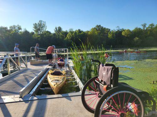 An empty manual wheelchair rests on the BoardSafe accessible launch dock while its user paddles out onto the lake, with more kayakers gliding across the green water beyond the cattails.