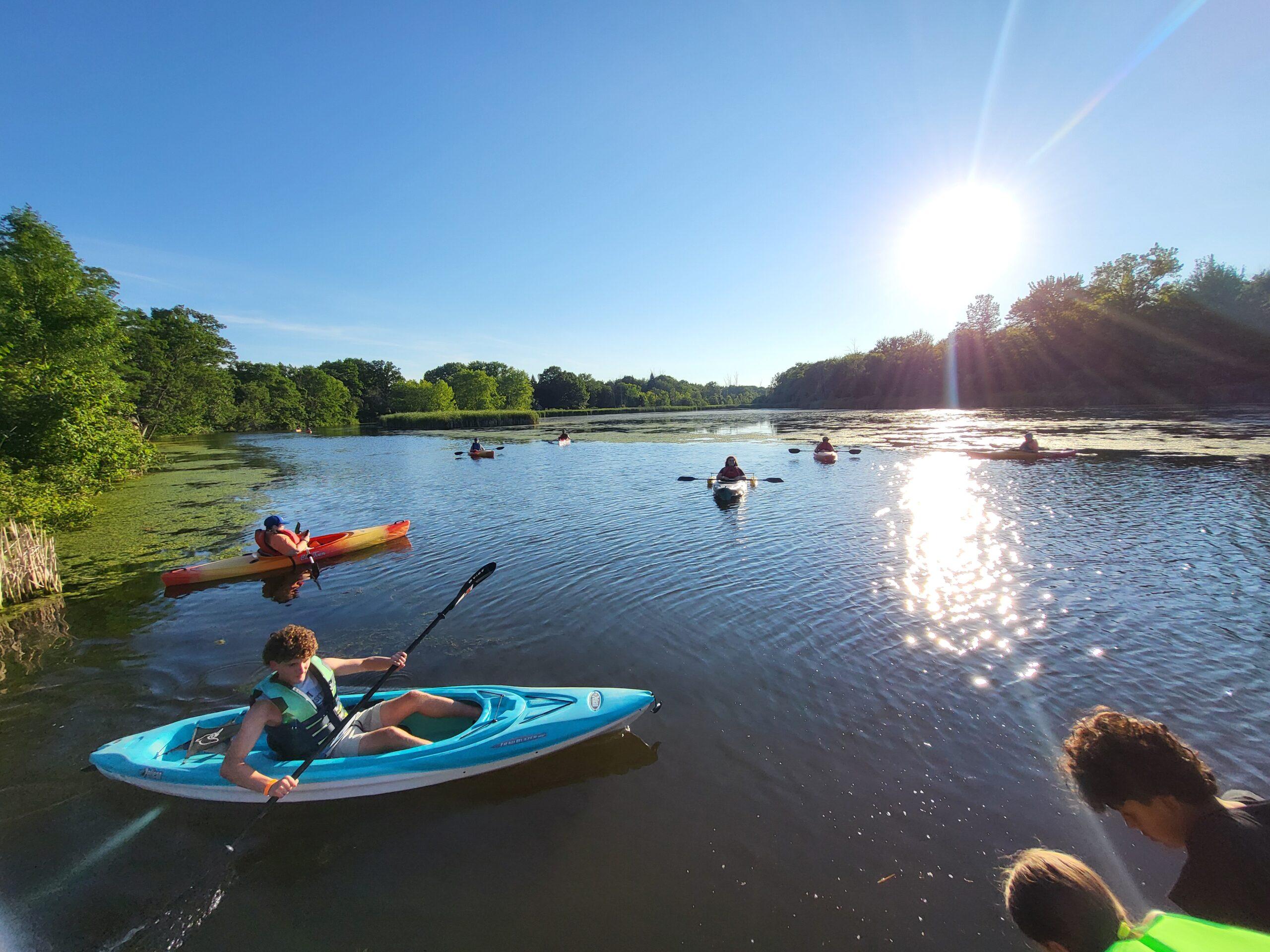 Several paddlers spread across a calm, sunlit lake in the early evening, with a young person in a blue kayak paddling in the foreground.