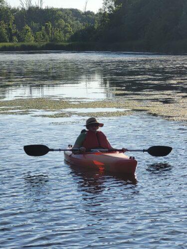 A woman sits peacefully in a kayak on a calm river late in the evening surrounded by water and trees